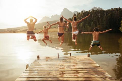 Fünf junge Freude springen von einem Steg in den See. Es ist ein schöner, sonniger Tag.
