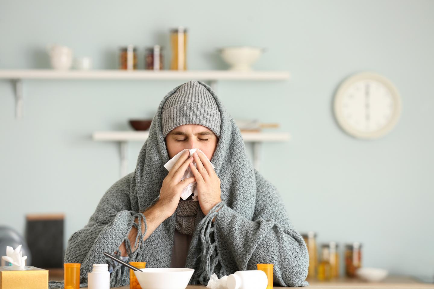 Ein kranker junger Mann sitzt in eine Decke gehüllt an einem Küchentisch und putzt sich die Nase