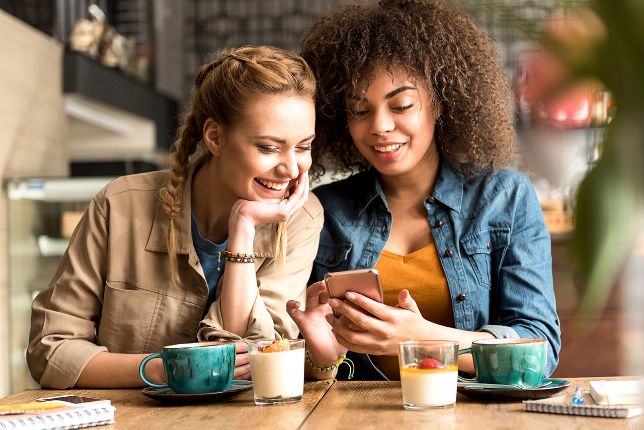 Outgoing girl speaking with cheerful african friend while sitting at table in confectionary shop. They looking at mobile