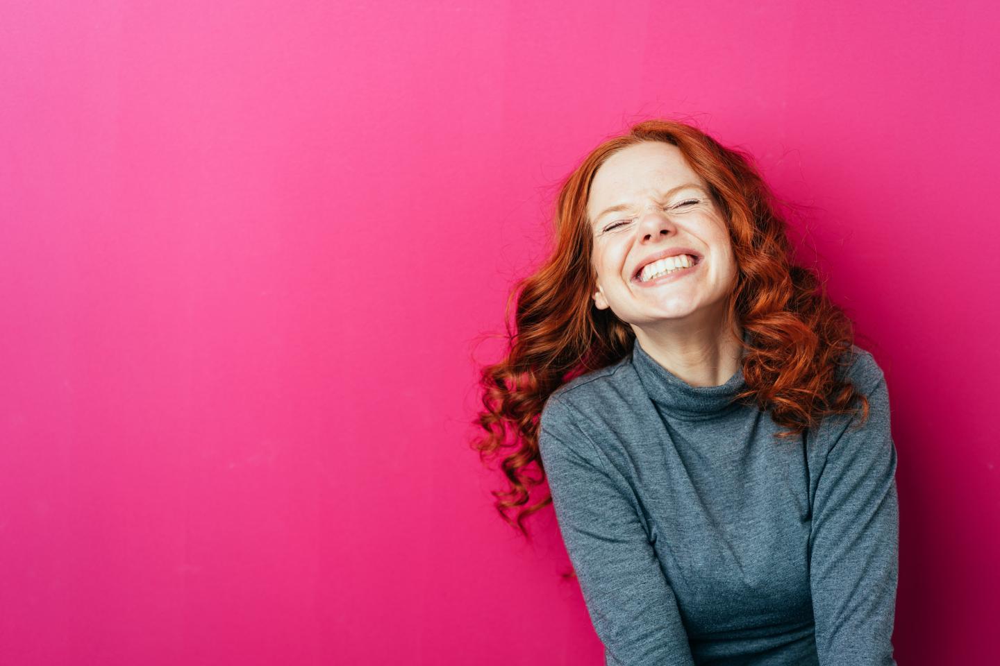 Junge Frau mit roten Haaren strahlt vor pinkfarbenem Hintergrund.