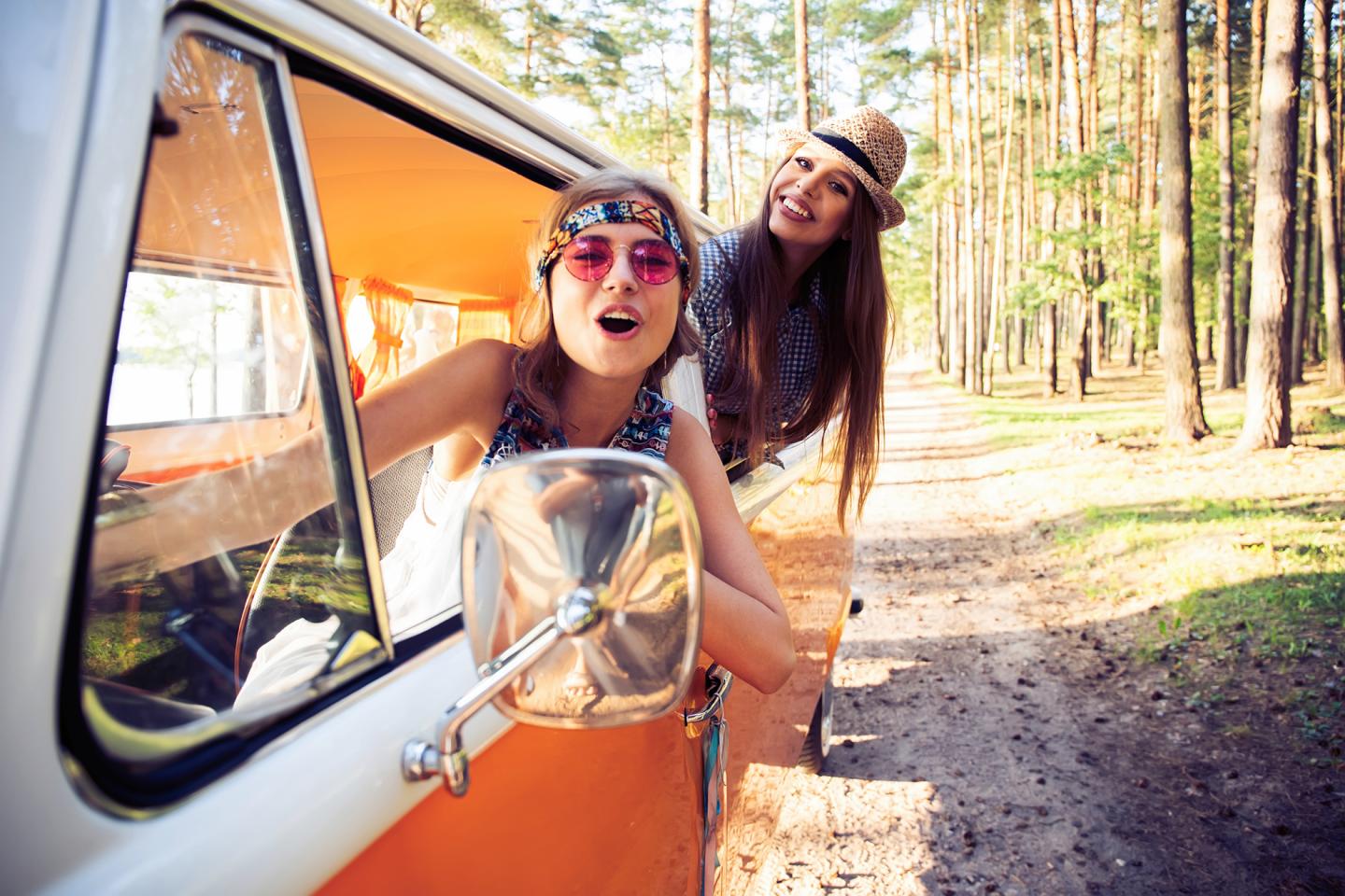Zwei Freundinnen fahren an einem sonnigen Tag in einem Kleinbus einen Waldweg entlang. Sie lachen glücklich in die Kamera.