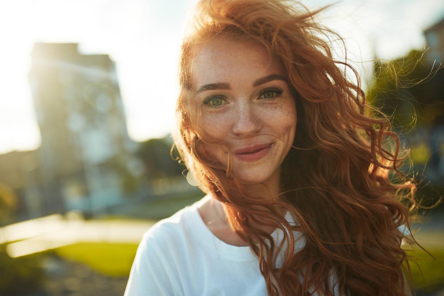 Portrait einer jungen Frau. Sie hat lockige rote Haare, blickt lächelnd in die Kamera.