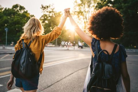 Zwei Freundinnen laufen Richtung Sonnenuntergang. Sie heben die Arme und halten sich dabei an der Hand.