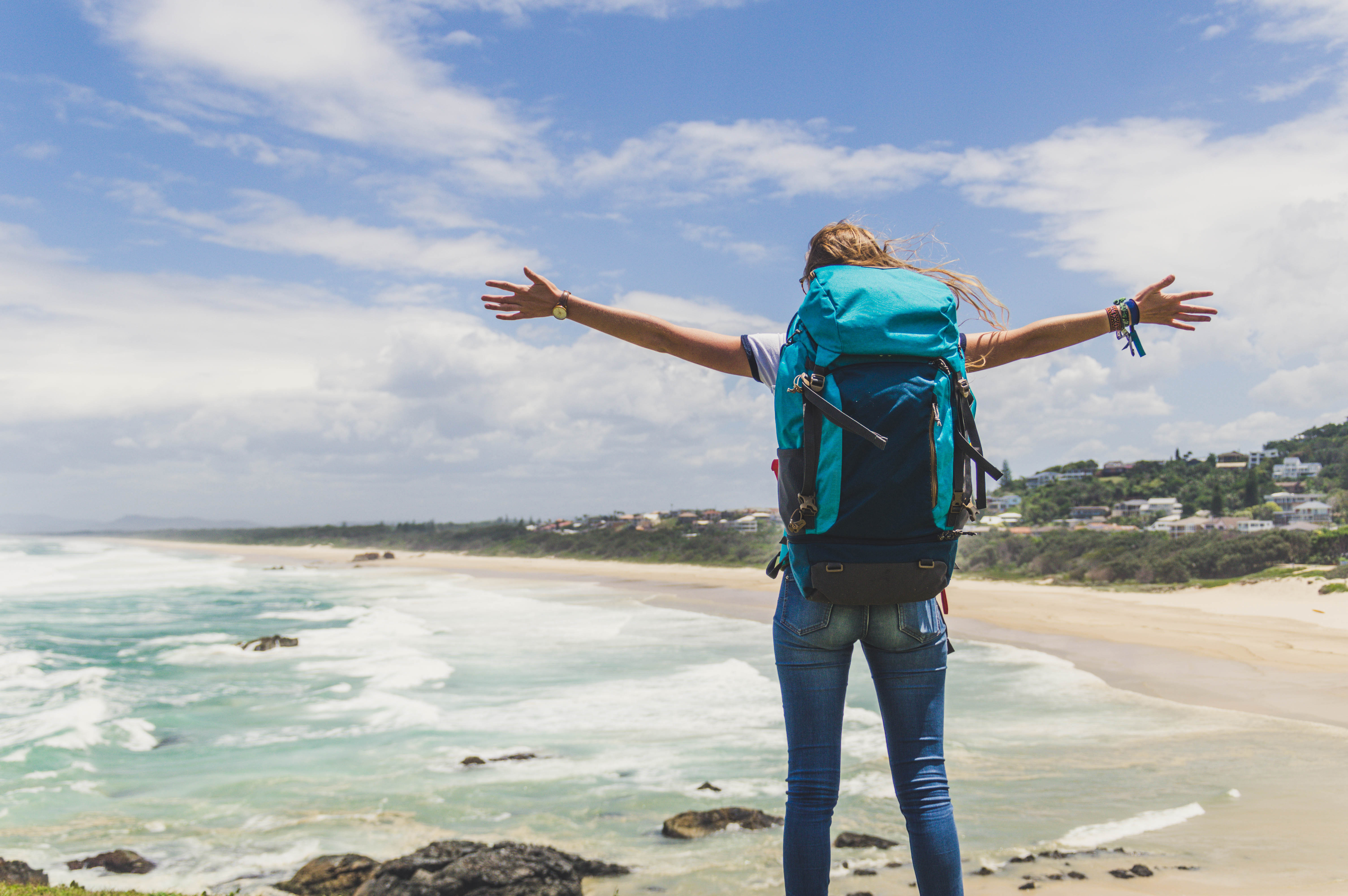 Eine Backpackerin breitet grüßend ihre Arme vor einer Landschaft aus. Sie zeigt einen Strand, im Hintergrund Bäume und Häuser.