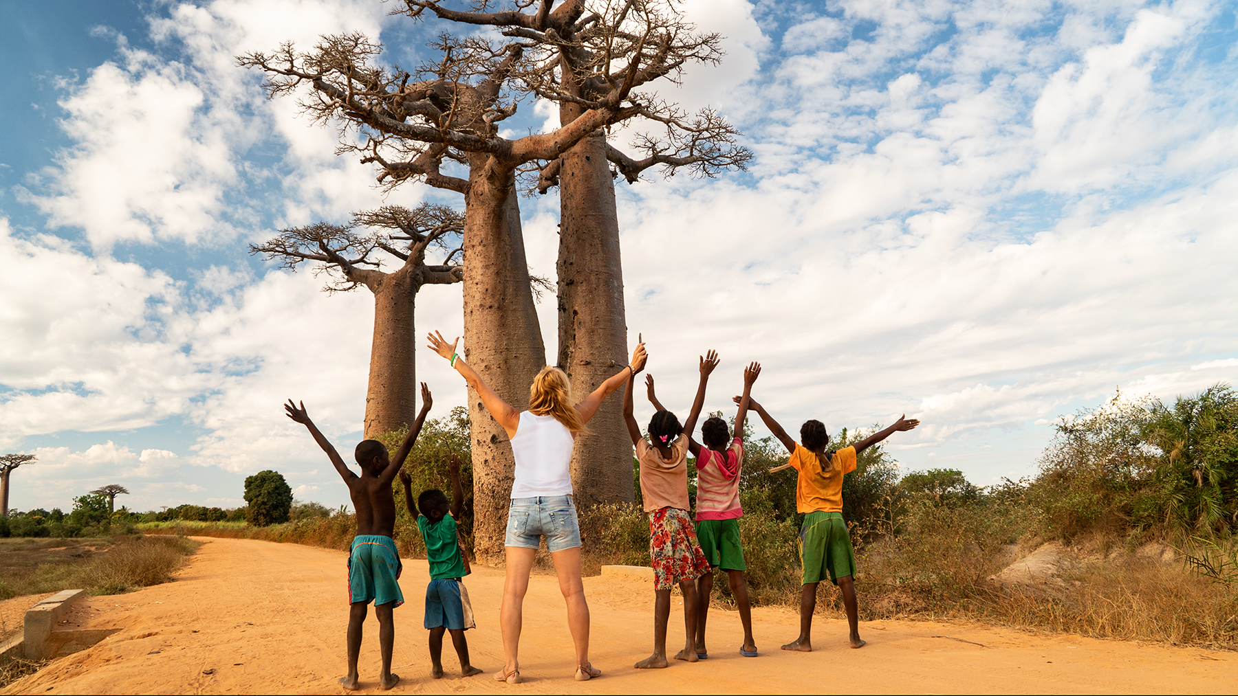 Fünf Kinder und eine erwachsene Frau heben ihre Arme in die Luft. Sie stehen vor 2 Afrikanischen Affenbäumen mit dem Rücken zur Kamera.