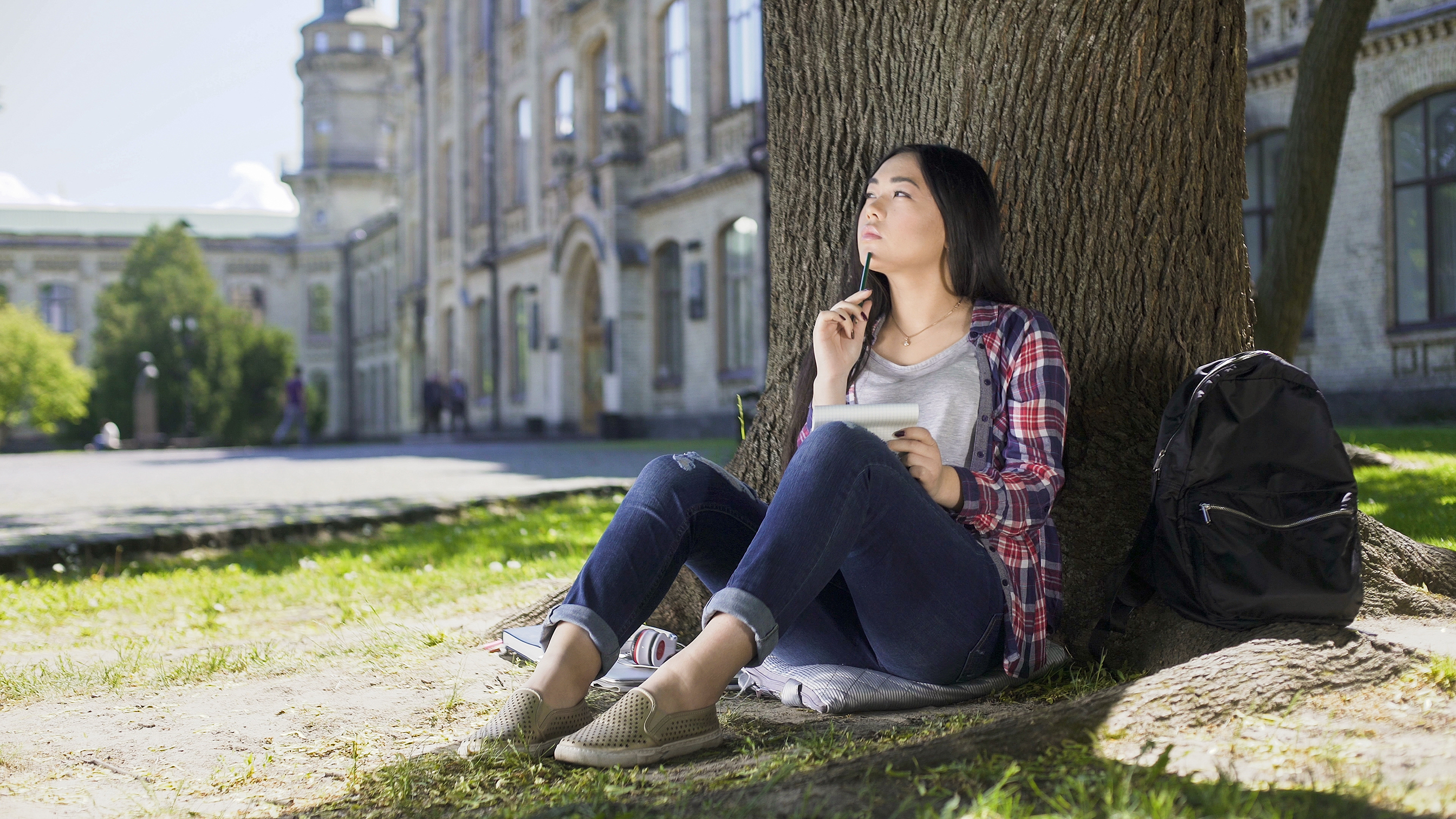 Eine Studentin sitzt im Gras und lehnt sich nachdenklich an einen Baum. Sie befindet sich auf einem Universitätscampus und hält Block und Stift.