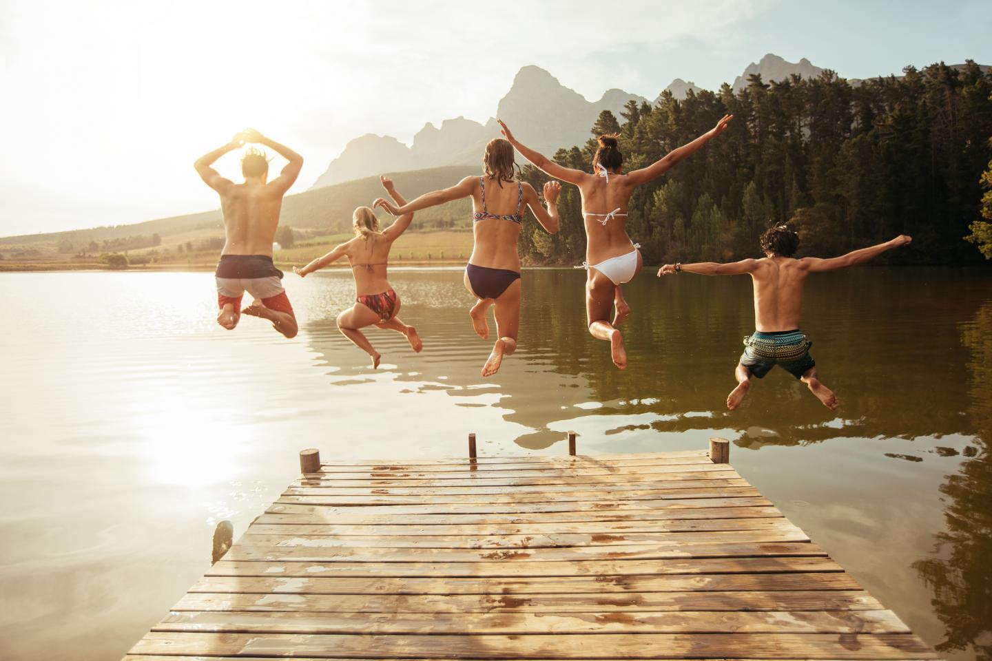 Fünf junge Freude springen von einem Steg in den See. Es ist ein schöner, sonniger Tag.