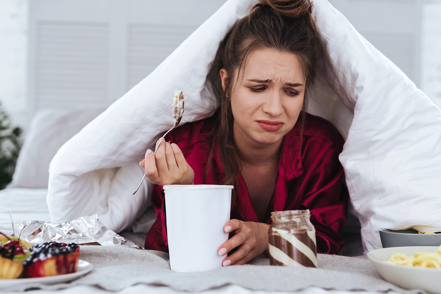 Eine junge Frau isst Eis im Bett. Sie ist umgeben von Süßigkeiten.