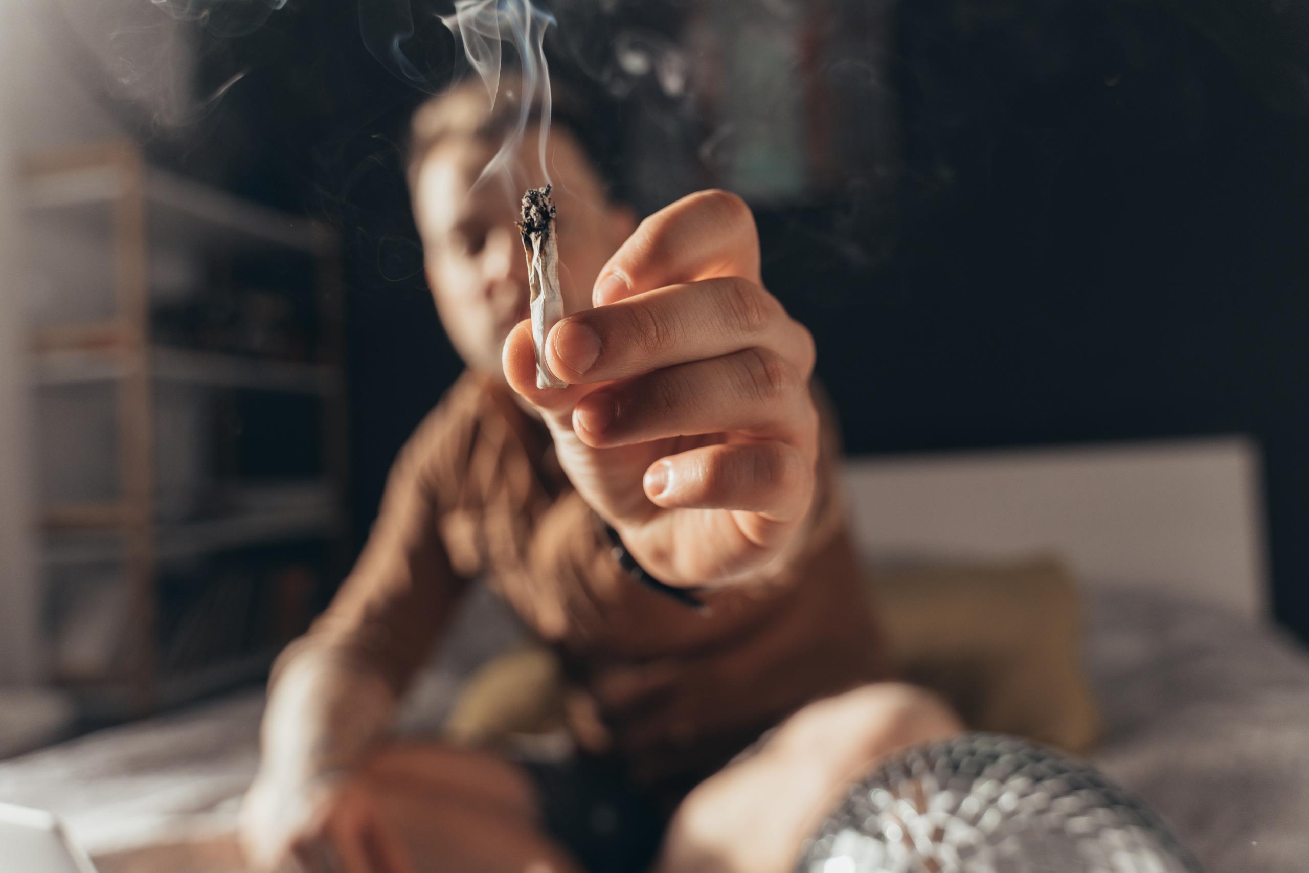 Take it. Close up view of the bearded man offering a hand rolled cannabis cigarette while sitting at the bed indoors. Focus at the cigarette with smoke