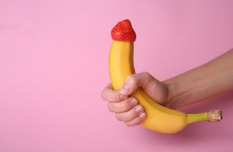 Hand hält Banane mit Erdbeere auf der Spitze vor rosa Hintergrund