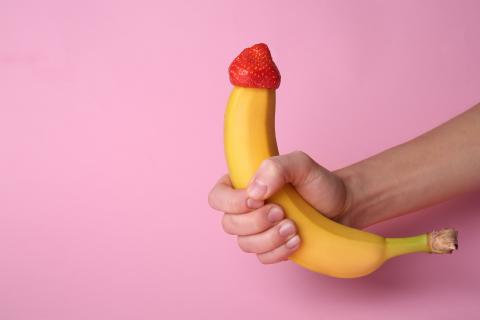 Hand hält Banane mit Erdbeere auf der Spitze vor rosa Hintergrund