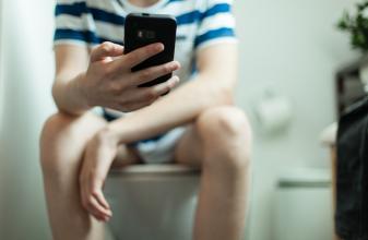 Boy sitting on a toilet using his mobile phone to text messages to his friends as he does his ablutions, close up on his hand