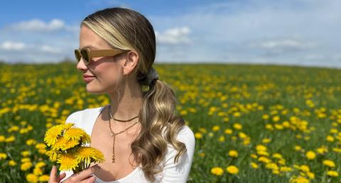 Frau mit blondem Zopf und Sonnebrille sitzt mit einem Strauß in der Hand in einem Feld mit Löwenzahn.